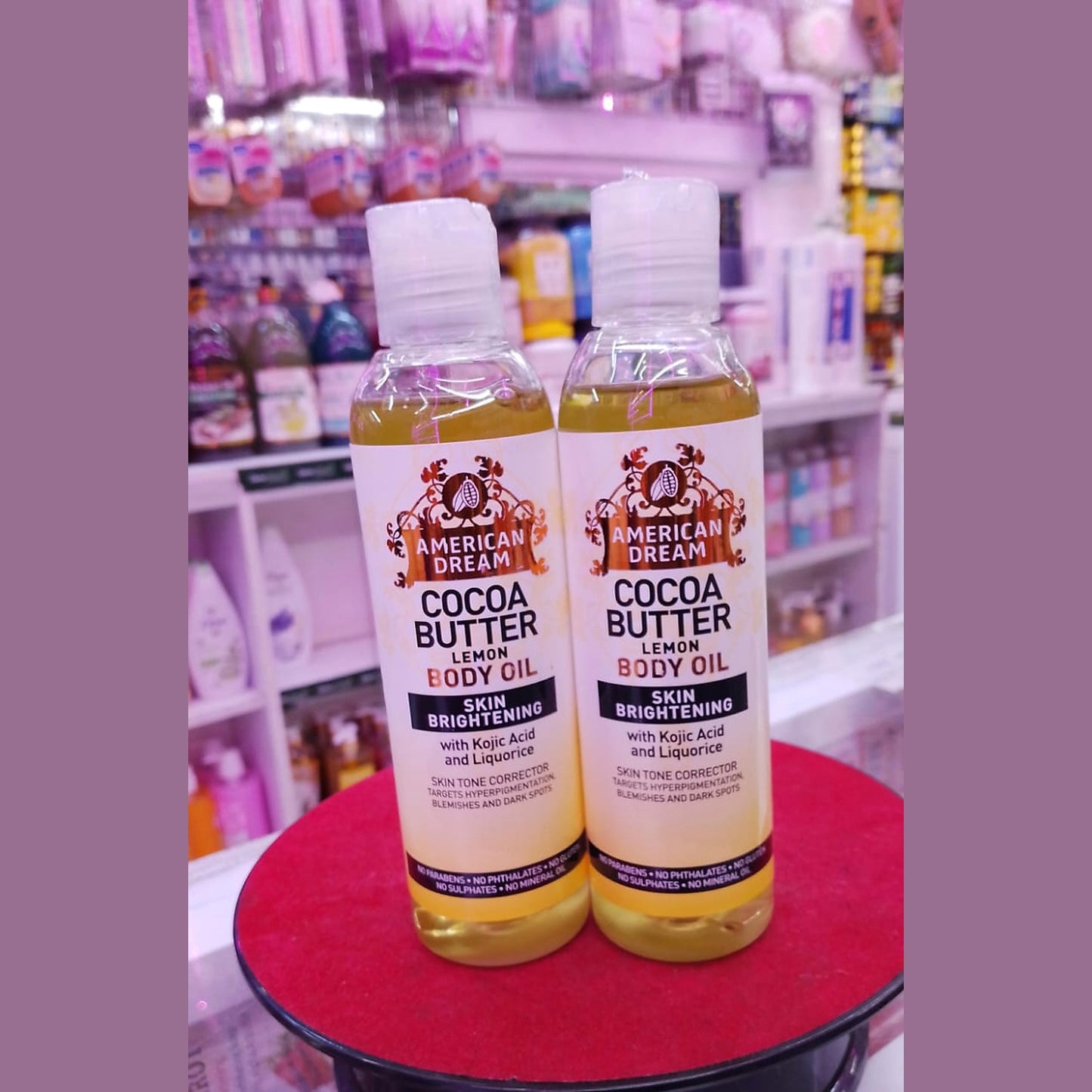 Two bottles of American Dream Cocoa Butter Lemon Body Oil on a red surface with a store shelf in the background.