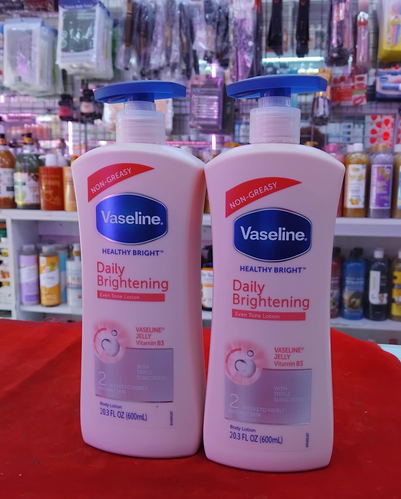 Two bottles of Vaseline Healthy Bright Daily Brightening lotion on a red surface with a store shelf in the background.