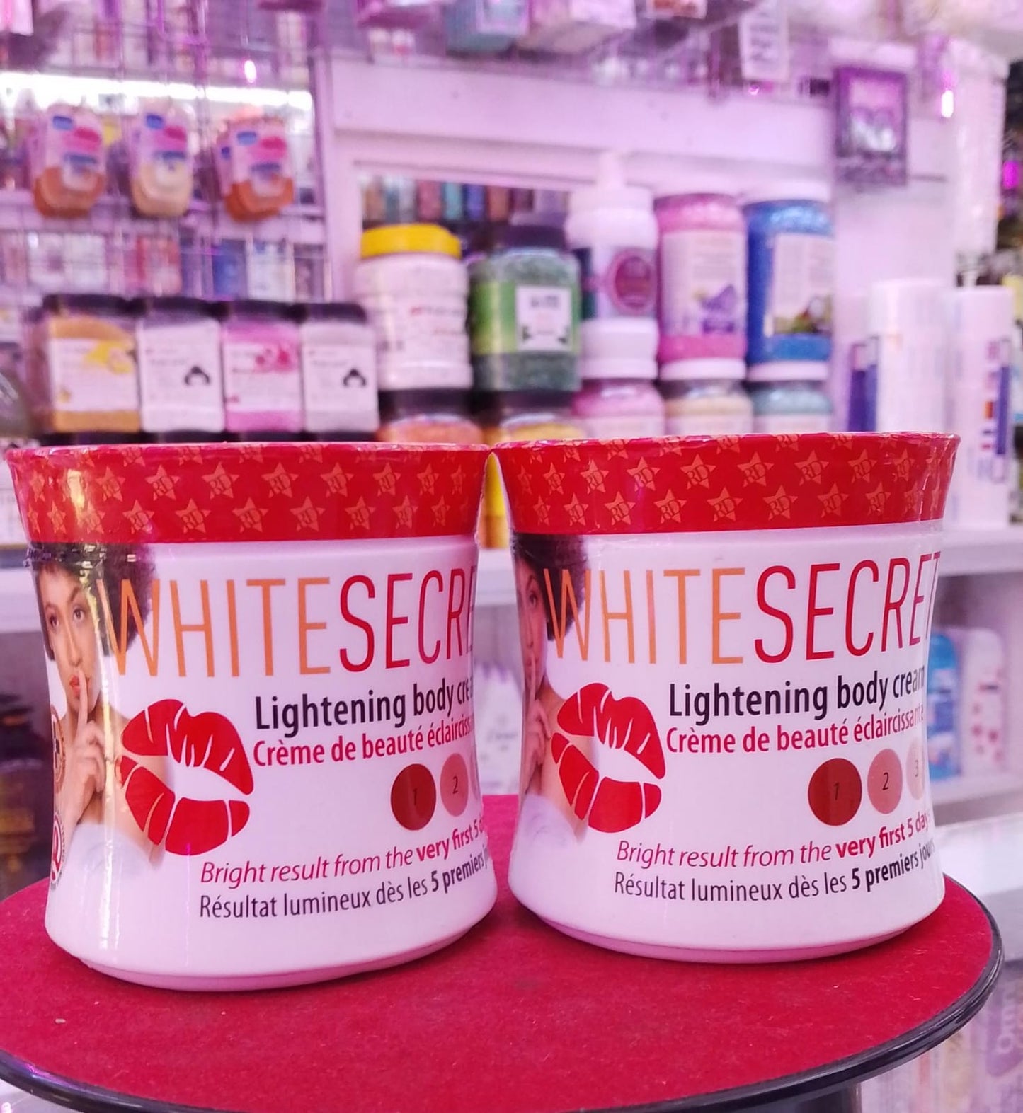 Two containers of 'White Secret' lightening body cream on a red surface with a blurred store background.