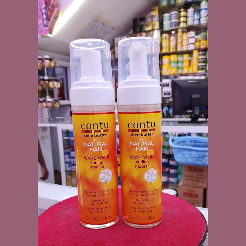 Two bottles of Cantu Natural Hair Wave Whip curling mousse on a red surface with a store shelf in the background.