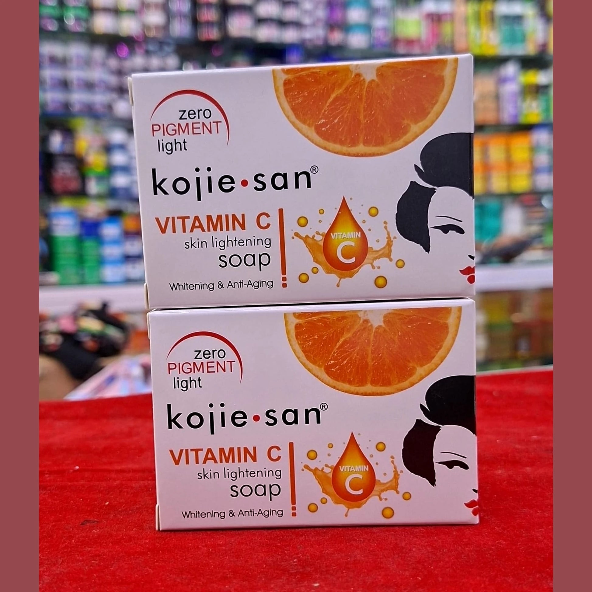 Two boxes of Kojie San Vitamin C soap on a red surface with a blurred store background.